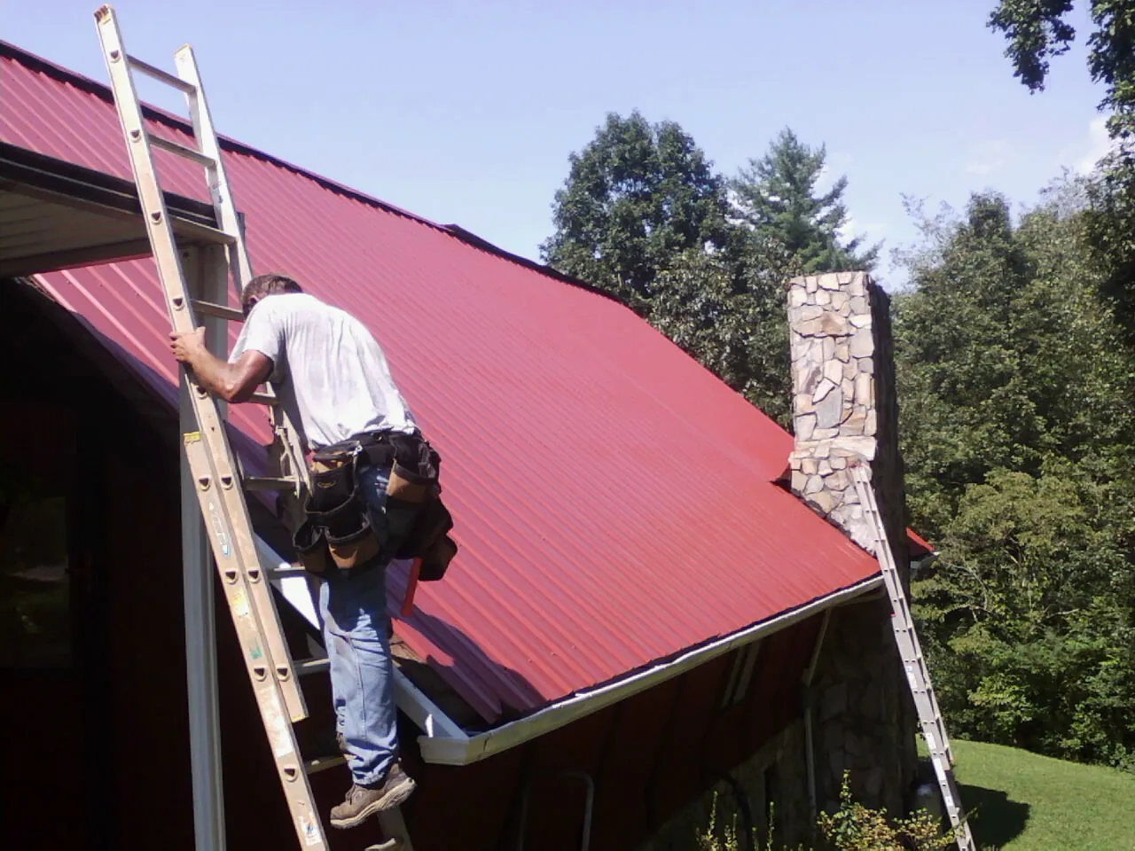 Roofer on a ladder beside a red metal roof and stone chimney — Soffit & Fascia Repair by Roofing Brothers in Oakville